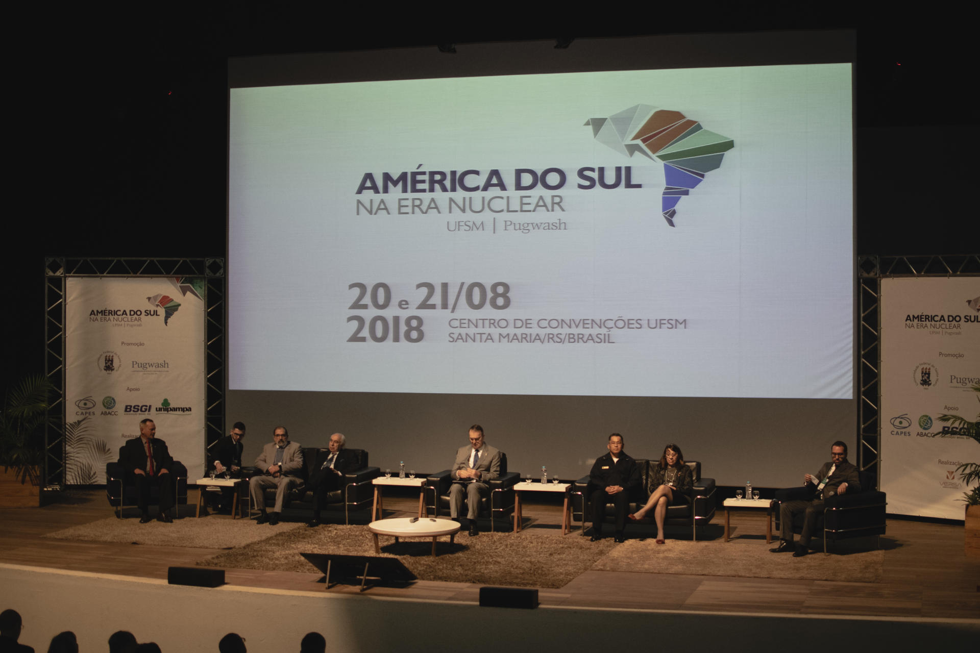 Foto horizontal colorida de um palco com pessoas sentadas em cadeiras. Ao fundo, um grande painel escrito América do Sul na Era Nuclear, 20 e 21/08 2018