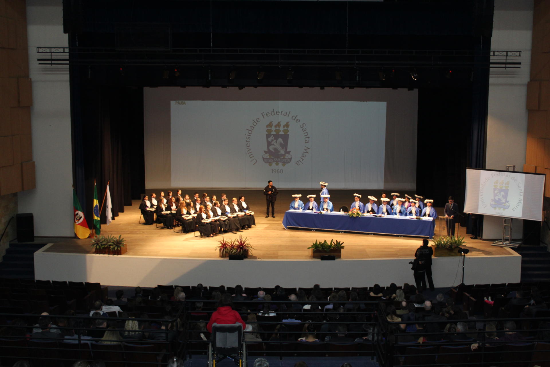 Imagem horizontal colorida vista de frente, à distância, de formandos em um palco