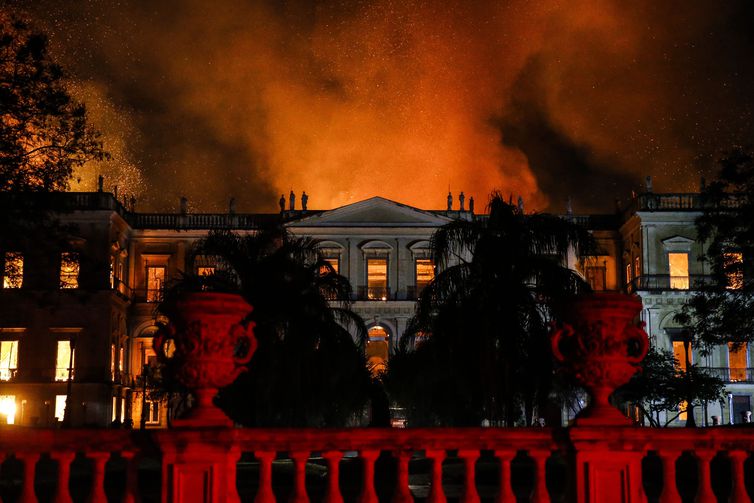 Fotografia com imagem em primeiro plano do muro do museu em vermelho e, em segundo plano, o prédio do museu em chamas. É possível perceber bombeiros em cima do prédio tentando controlar o fogo.