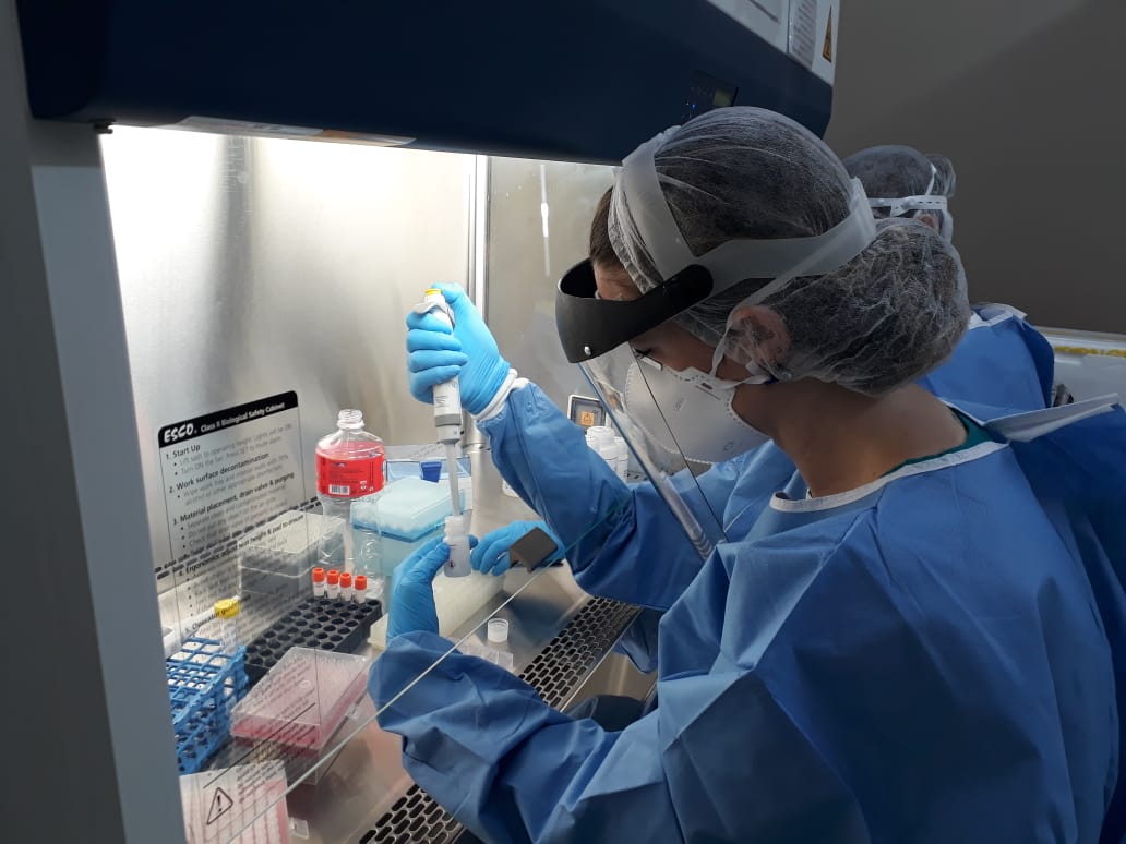 Dois profissionais usando equipamentos de proteção trabalhando em uma bancada com seringas e materiais hospitalares