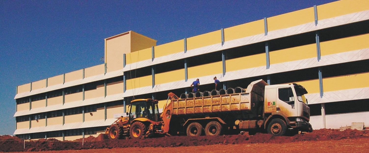 Foto horizontal colorida de prédio de 4 andares amarelo com branco e azul, na frente chão de terra vermelha, um trator e um caminhão com 2 trabalhadores em cima
