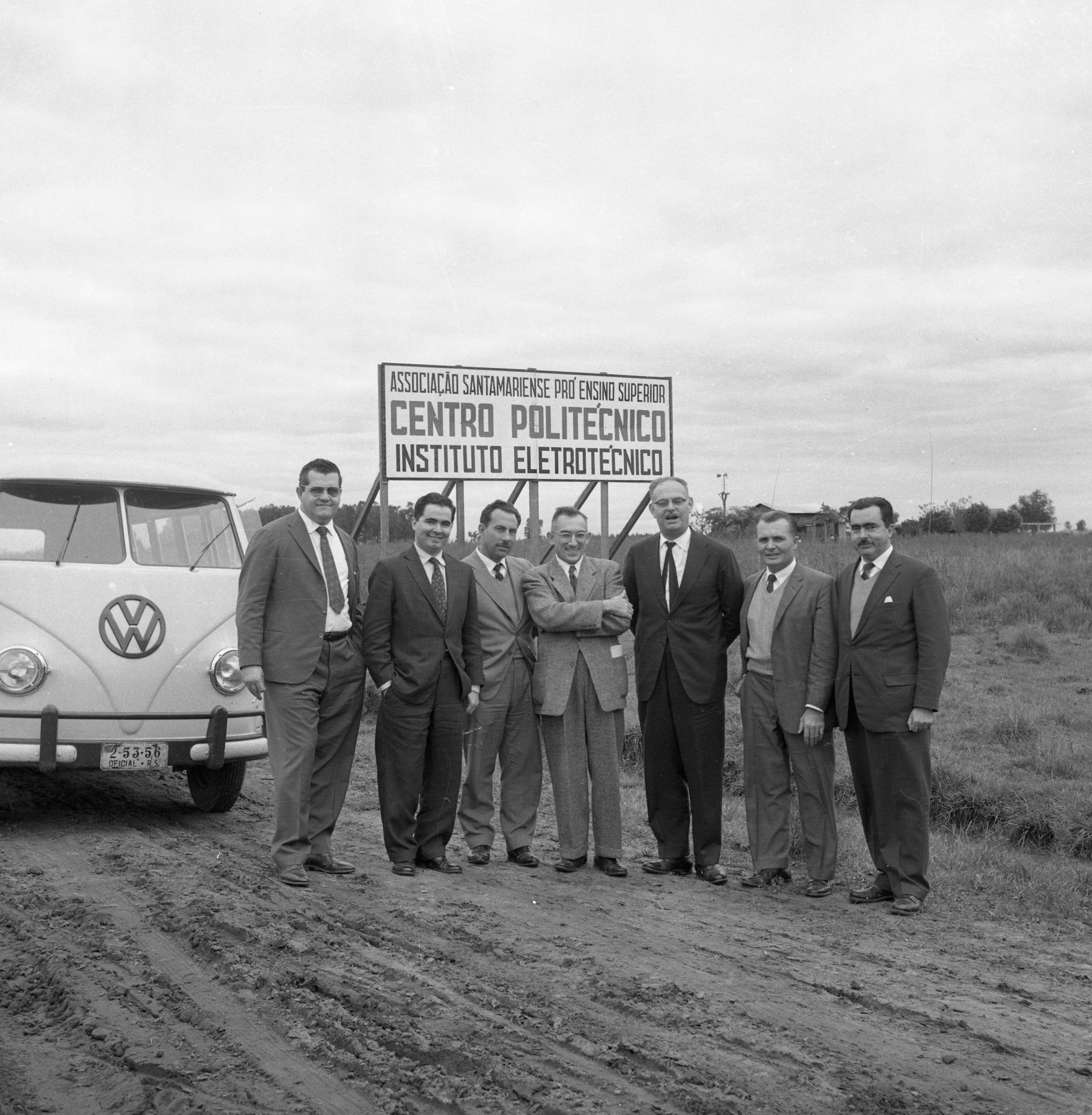 Fotografia quadrada preto e branca de 7 homens em uma estrada de chão em frente de uma Kombi e de uma placa escrita "Associação Santamariense Pró-Ensino Superior - Centro Politécnico - Instituto Eletrotécnico".
