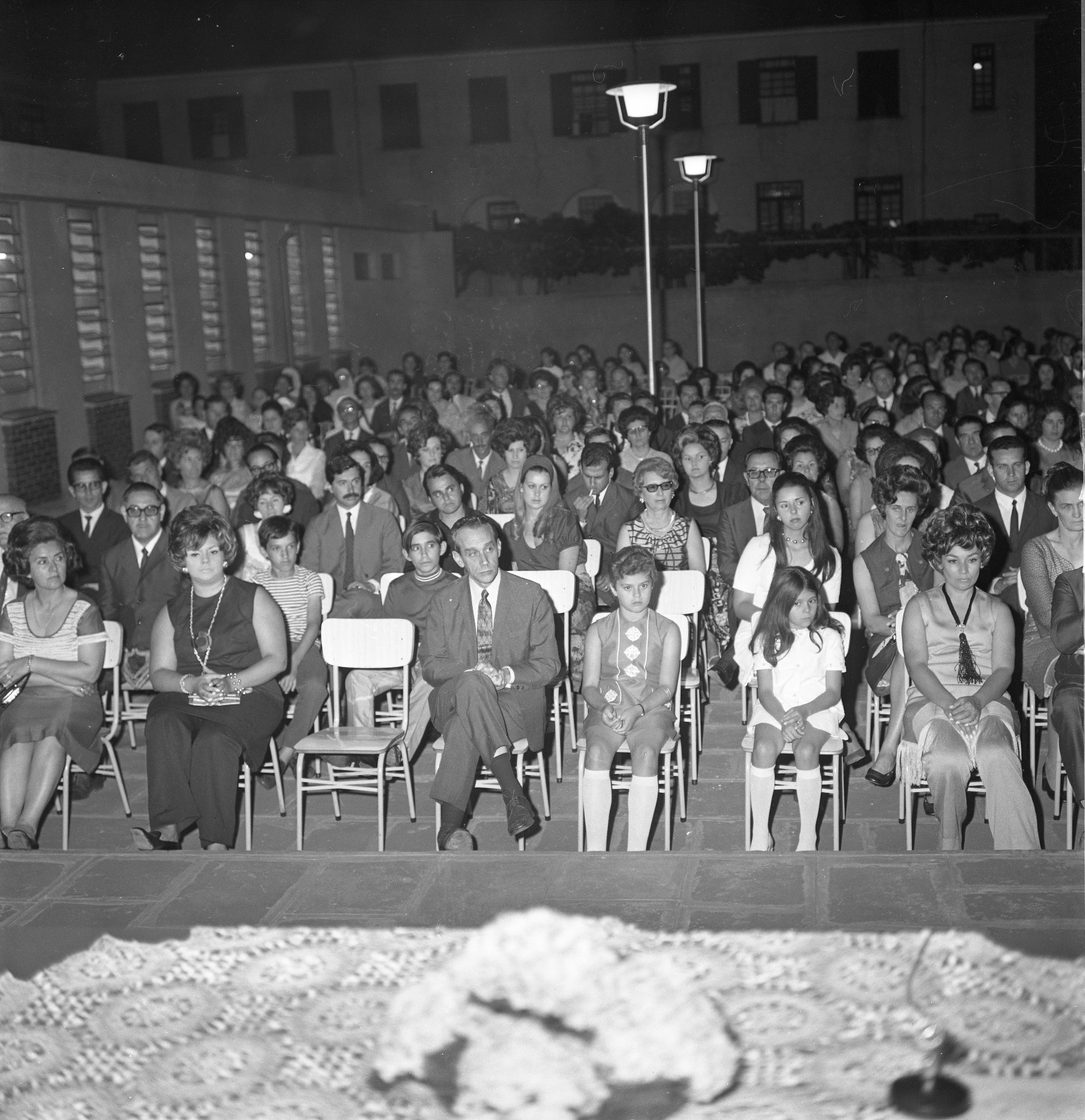 Fotografia quadrada preto e branca da inauguração de um pavilhão da Extensão da UFSM em São Borja. A fotografia foi tirada de cima de um palco, de onde vê-se o público, que está que está sentado um nível abaixo em 9 fileiras de cadeiras escolares.