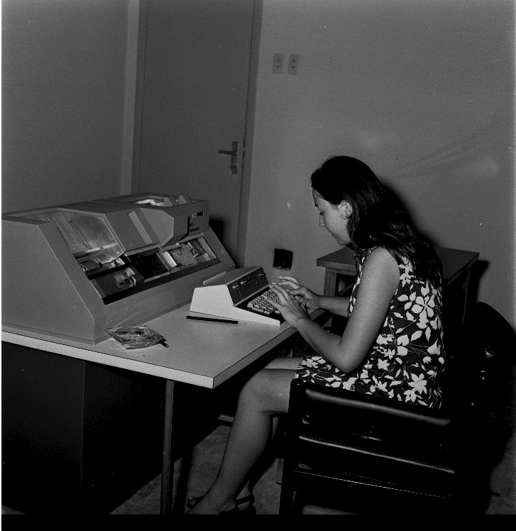 Fotografia quadrada preto e branca de uma mulher sentada a frente de um computador eletrônico em uma sala. A mulher é jovem, veste vestido florido e digita e olha em direção ao computador.