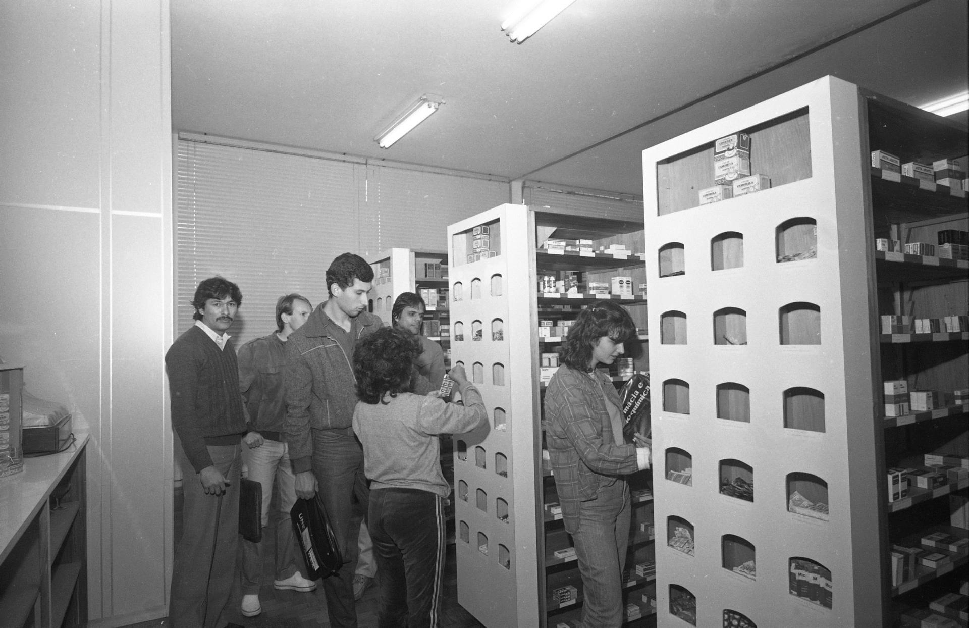 Fotografia horizontal preto e branca da Inauguração da Farmácia-escola. Em ambiente fechado, 6 pessoas estão próximas a 3 estantes com caixas de remédios expostos. Uma delas manipula uma cartela de comprimidos.