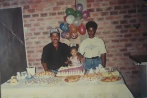 Foto colorida de três pessoas em uma sala. Uma criança ao centro, com um homem do lado direito e uma mulher do lado esquerda. Na frente deles, uma mesa com comidas e um bolo de aniversário.