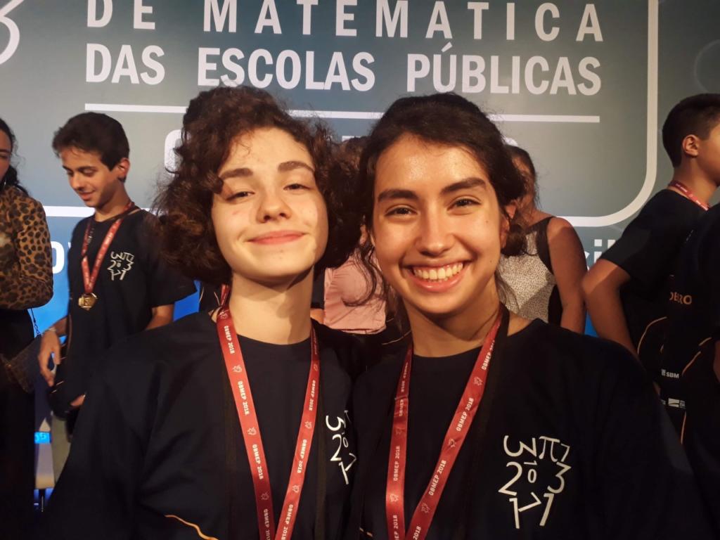 Foto colorida com duas meninas em destaque. As duas estão sorrindo e com medalhas no pescoço.
