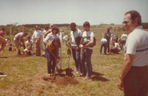 diversas pessoas em um campo aberto plantam mudas de árvores. Ao centro quatro alunos, duas meninas e dois meninos. Uma das meninas está vestida de amarelo e usa uma pá. Os demais estão vestidos de camisa branca e observam. No canto direito, em primeiro plano, um homem, grisalho, pele branca, óculos escuros, camisa branca e calça marrom. 