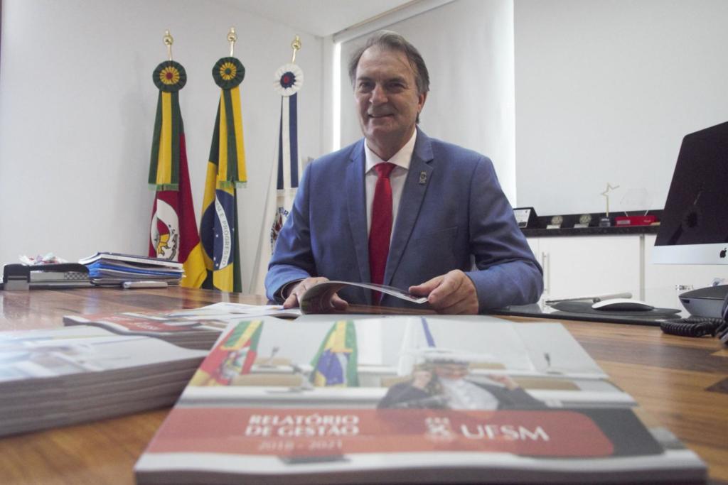 Foto colorida horizontal do reitor Paulo Burmann. Ele está sentado atrás da mesa. Em primeiro plano, é possível perceber o relatório de gestão. Ao fundo, as bandeiras da UFSM, do Rio Grande do Sul e do Brasil.