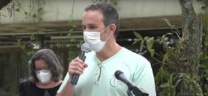 professor Fábio pacheco em primeiro plano, ao centro, fala ao microfone. Homem branco, cabelos curtos grisalhos, usa máscara e camiseta verde. Atrás dele está a cerimonialista do evento. 