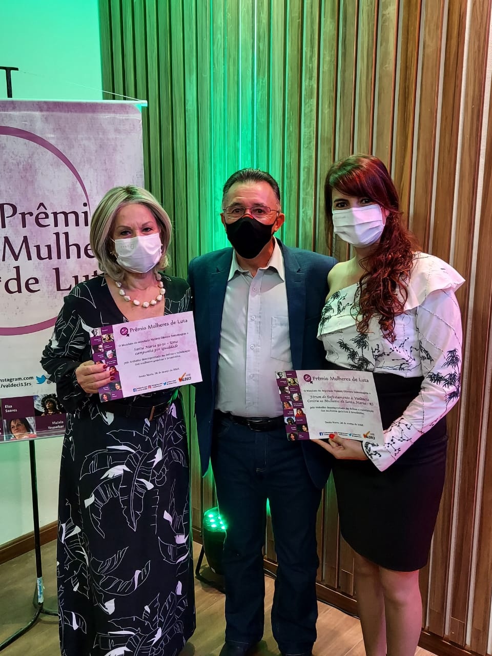 Foto vertical colorida mostra as duas professoras segurando seus certificados, ao centro o deputado, ao fundo um banner da premiação. Todos usam máscaras