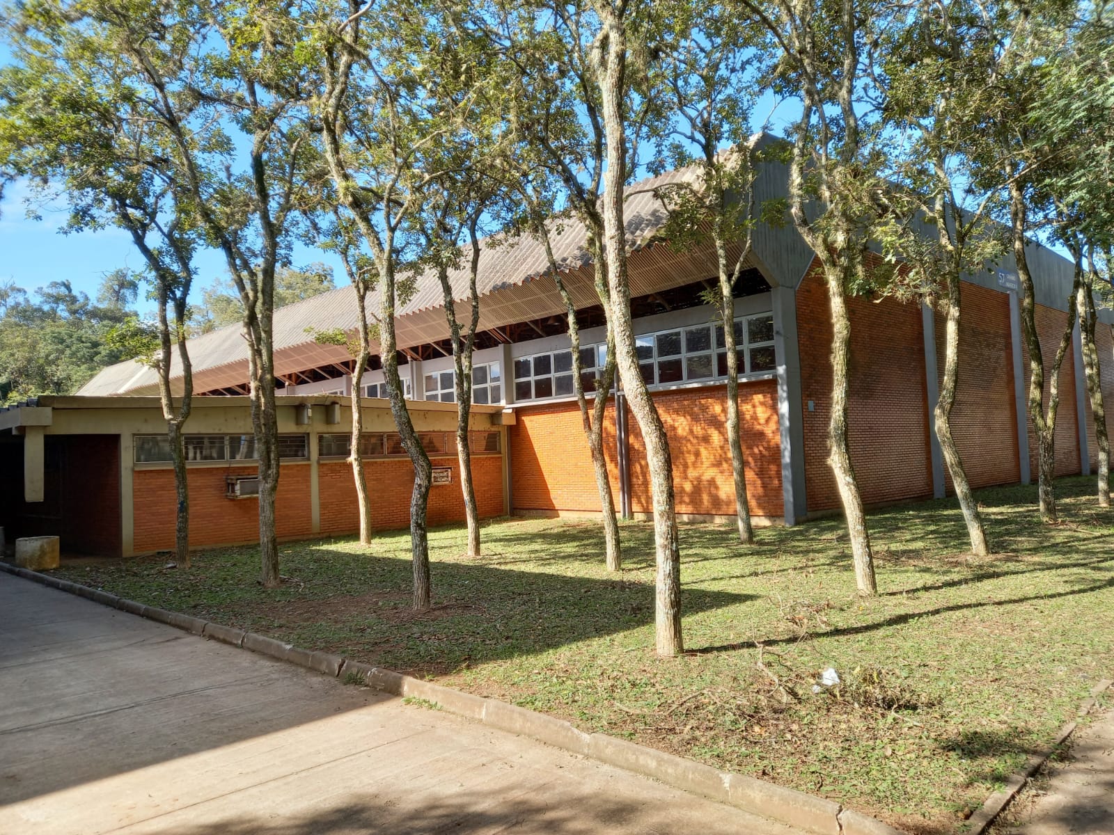 Foto colorida horizontal mostra a fachada do ginásio, com tijolos aparentes, e na frente um gramado com árvores em um dia de sol