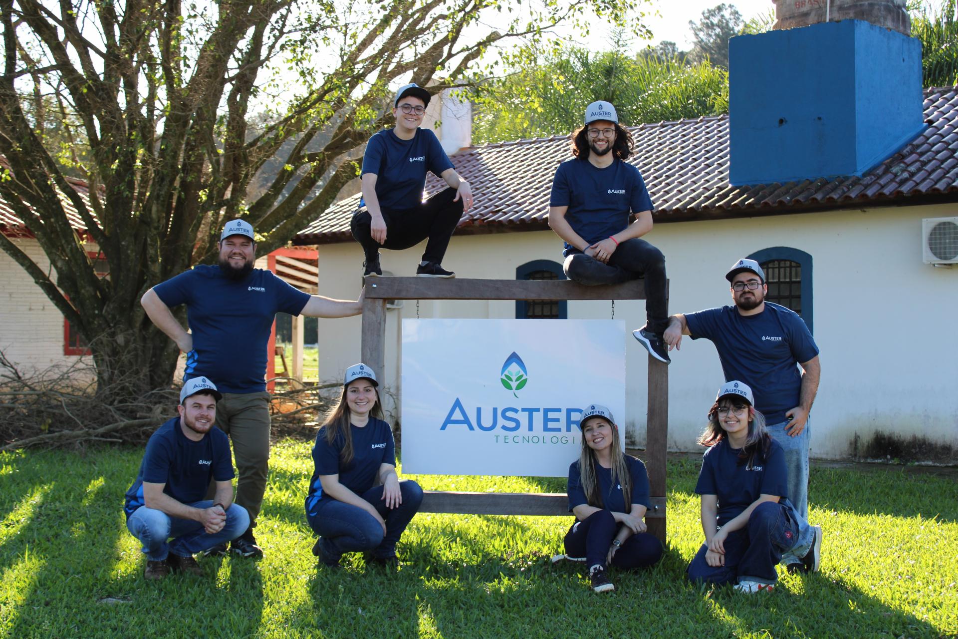 foto horizontal colorida mostra oito jovens, homens e mulheres, com camisetas azul marinho, ao redor de uma placa da Auster instalada em um gramado