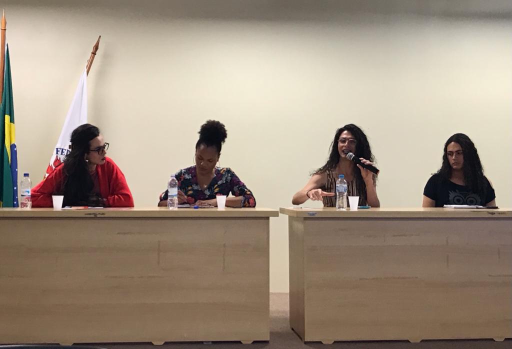 Foto horizontal de 4 mulheres sentadas. Elas estão atrás de uma mesa na cor marrom claro. A terceira mulheres, da esquerda para a direita, segura um microfone. Do lado esquerdo da foto, parte das bandeiras do Brasil e da UFSM. O fundo é bege.
