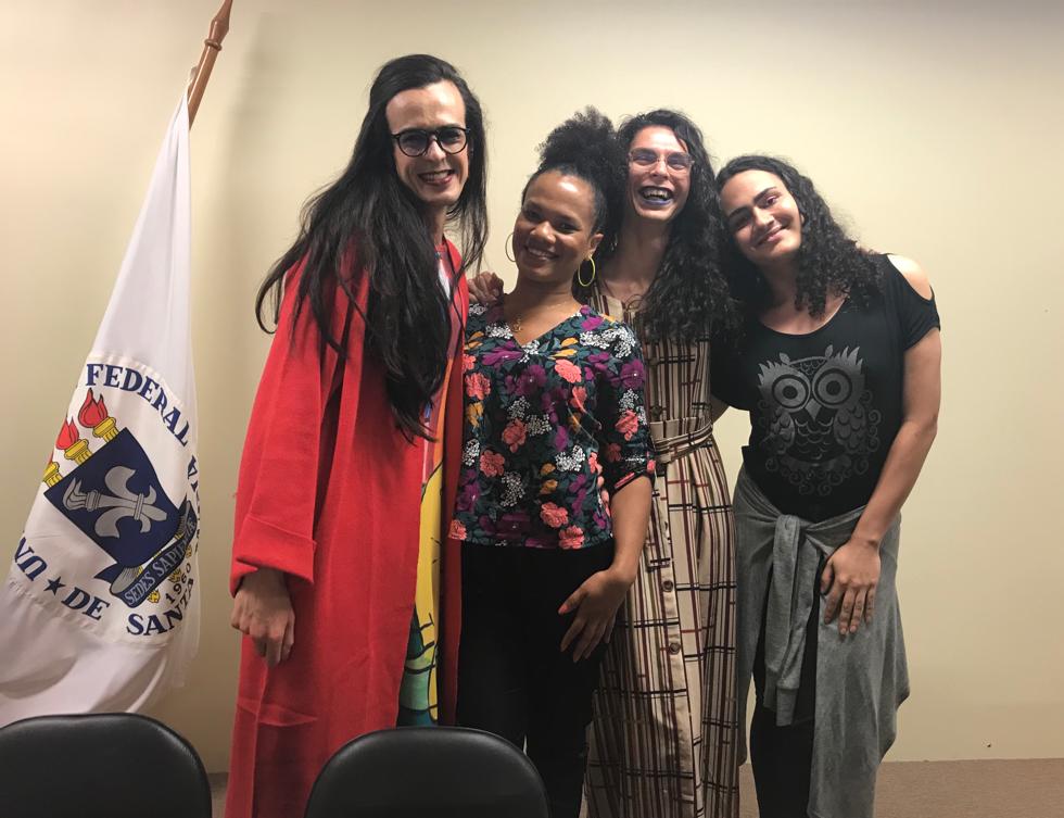 Foto horizontal de 4 mulheres abraçadas e sorrindo. Da esquerda para a direita estão Ali, Lauri, Rafa Ella e Raphaela. Do lado esquerdo da imagem, a bandeira da UFSM. Ao fundo uma parede na cor bege.
