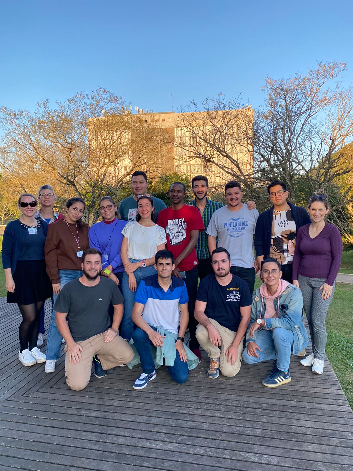 foto colorida vertical mostra jovens de ambos os sexos posando para a câmera em um deck de madeira do espaço multiuso, aparecendo ao fundo, ensolarado, o prédio da reitoria e árvores com galhos secos