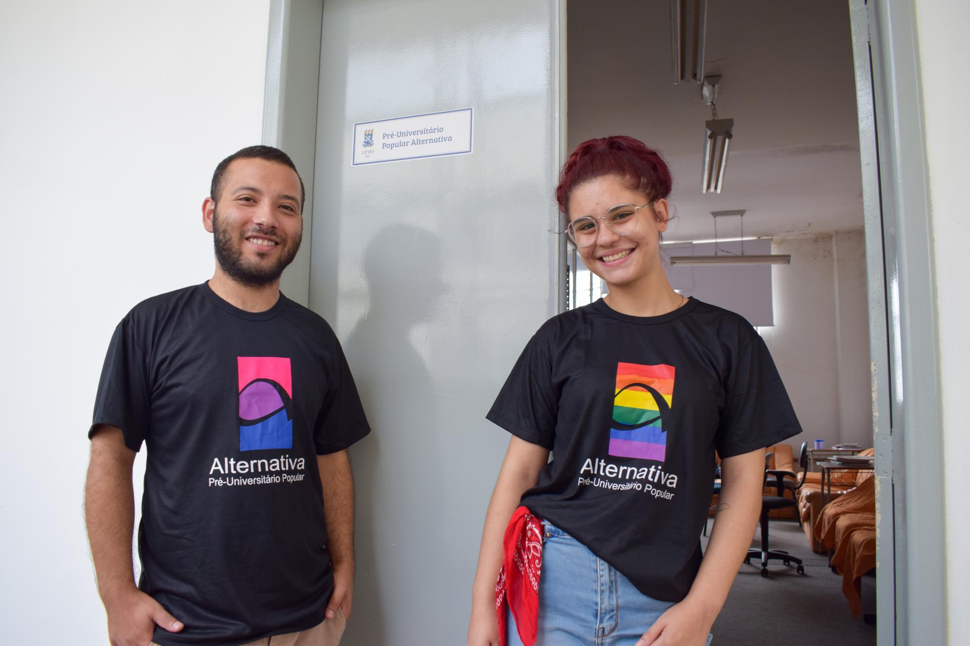foto colorida horizontal com duas pessoas, de frente, sorrindo, vestindo camisetas pretas com o logo colorido do Alternativa pré-vestibular