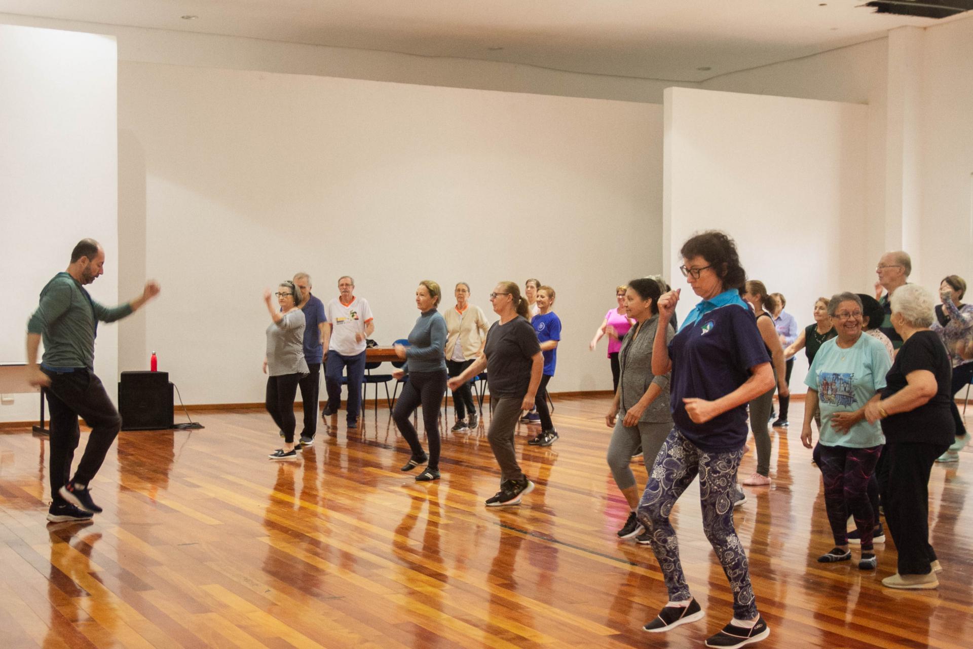 foto colorida horizontal com um grupo de pessoas, em sua maioria idosos, em uma sala ampla, fazendo movimentos de dança com os braços, e à frente delas, de frente para o grupo, um homem faz os movimentos que o grupo deve repetir