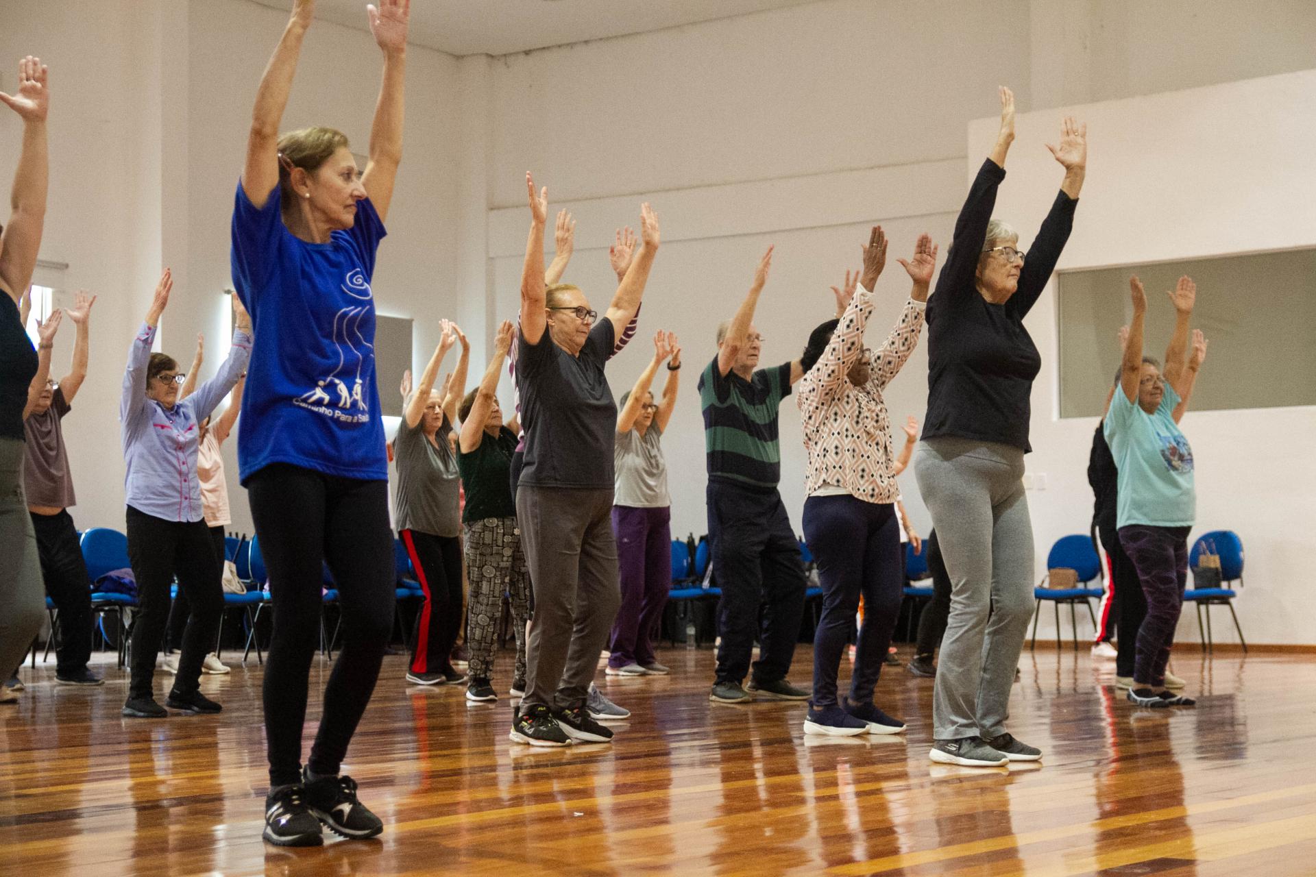 foto colorida horizontal com pessoas em um grande grupo se exercitando, em uma sala ampla com piso de madeira brilhoso. As pessoas estão com os dois braços para cima