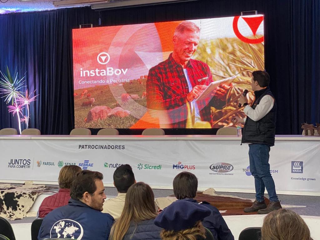 foto colorida horizontal com um palco, algumas pessoas de costas, um homem à frente virado para a tela, em que há um homem usando um tablet em uma lavoura