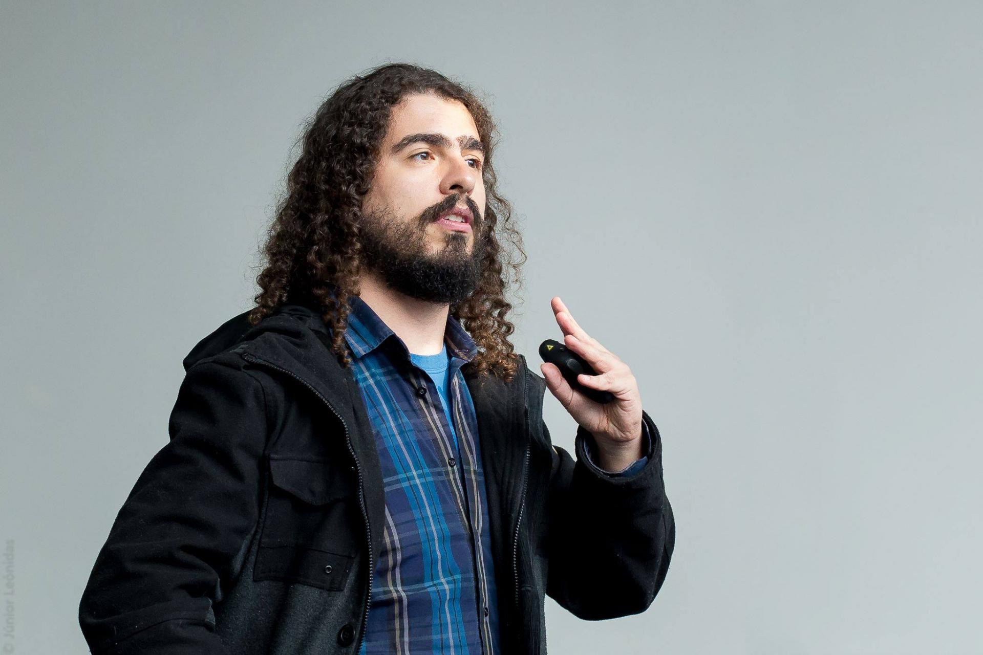 foto colorida de um homem de cabelo comprido, barba, vestido em tons escuros e azul, falando, atrás um fundo cinza