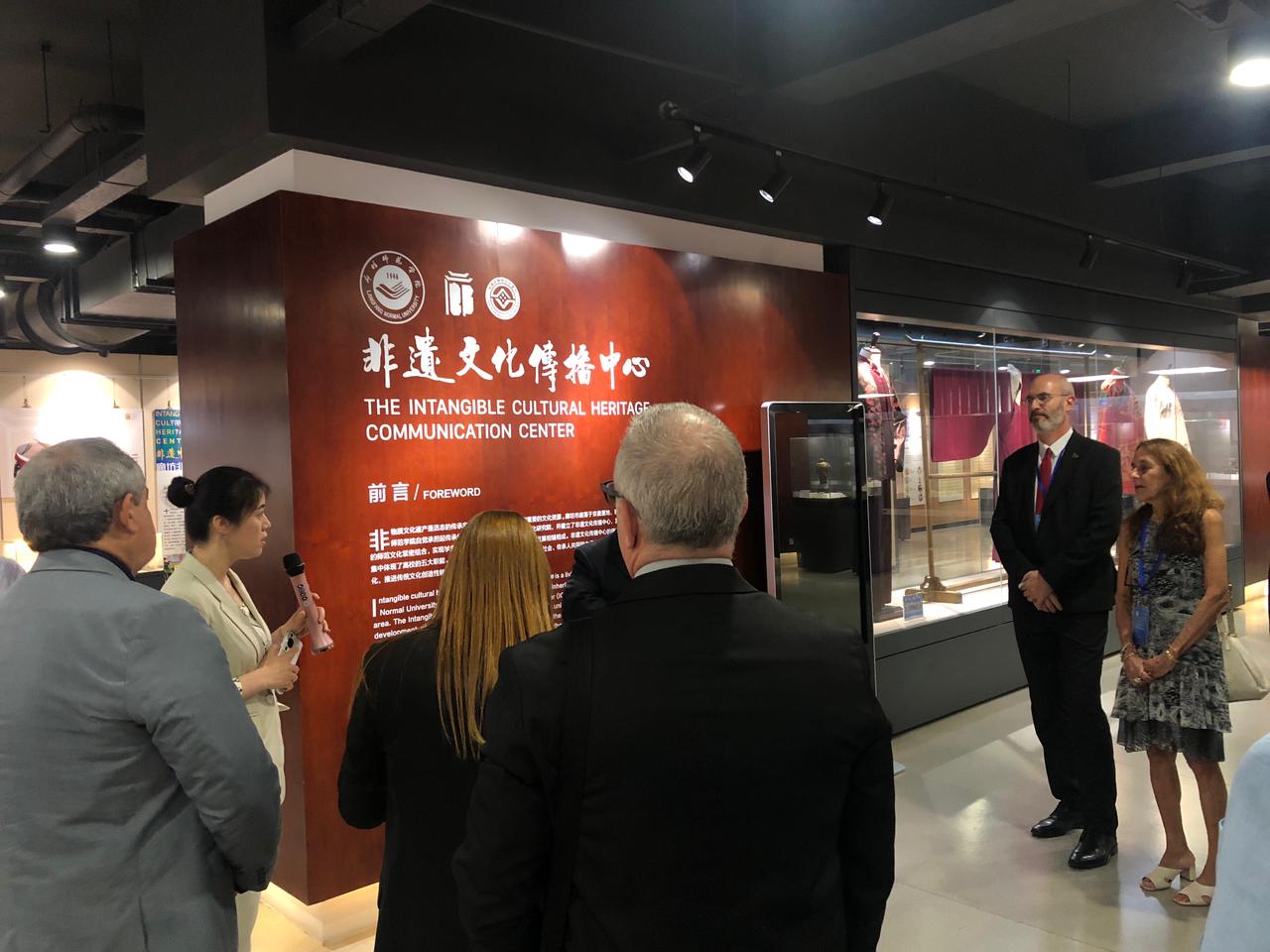 Na foto, várias pessoas em pé observam mulher com microfone que explica o que consta na área de exposição. Atrás da mulher um painel em vermelho com inscrições em branco em mandarim e em inglês.