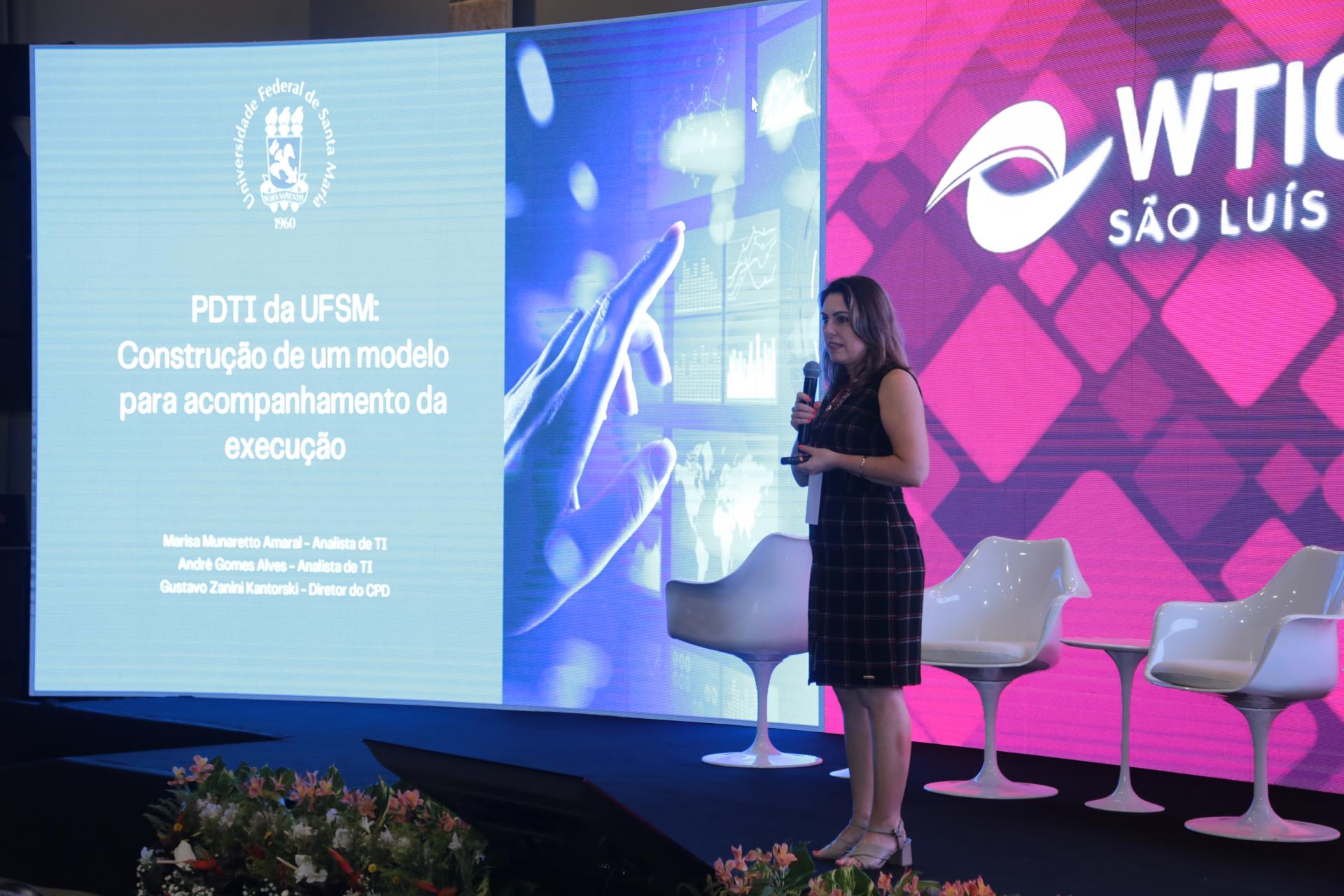 Mulher em pé no palco segura microfone. Atrás dela, um painel colorido