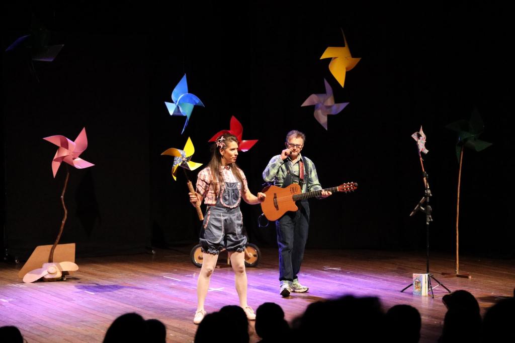 foto colorida horizontal com dois atores no palco, com luz baixa e cataventos coloridos pendentes do teto. O homem toca violão e a mulher segura um catavento