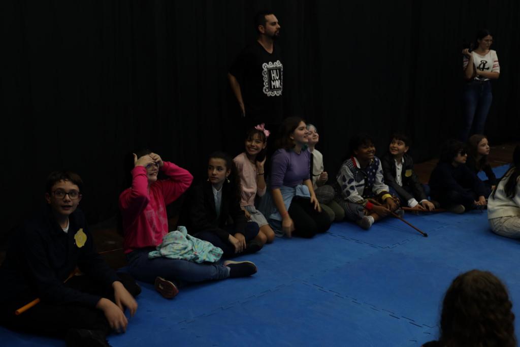 foto colorida horizontal com crianças sentadas num chão azul, em semicirculo, com alguns adultos atrás, em pé