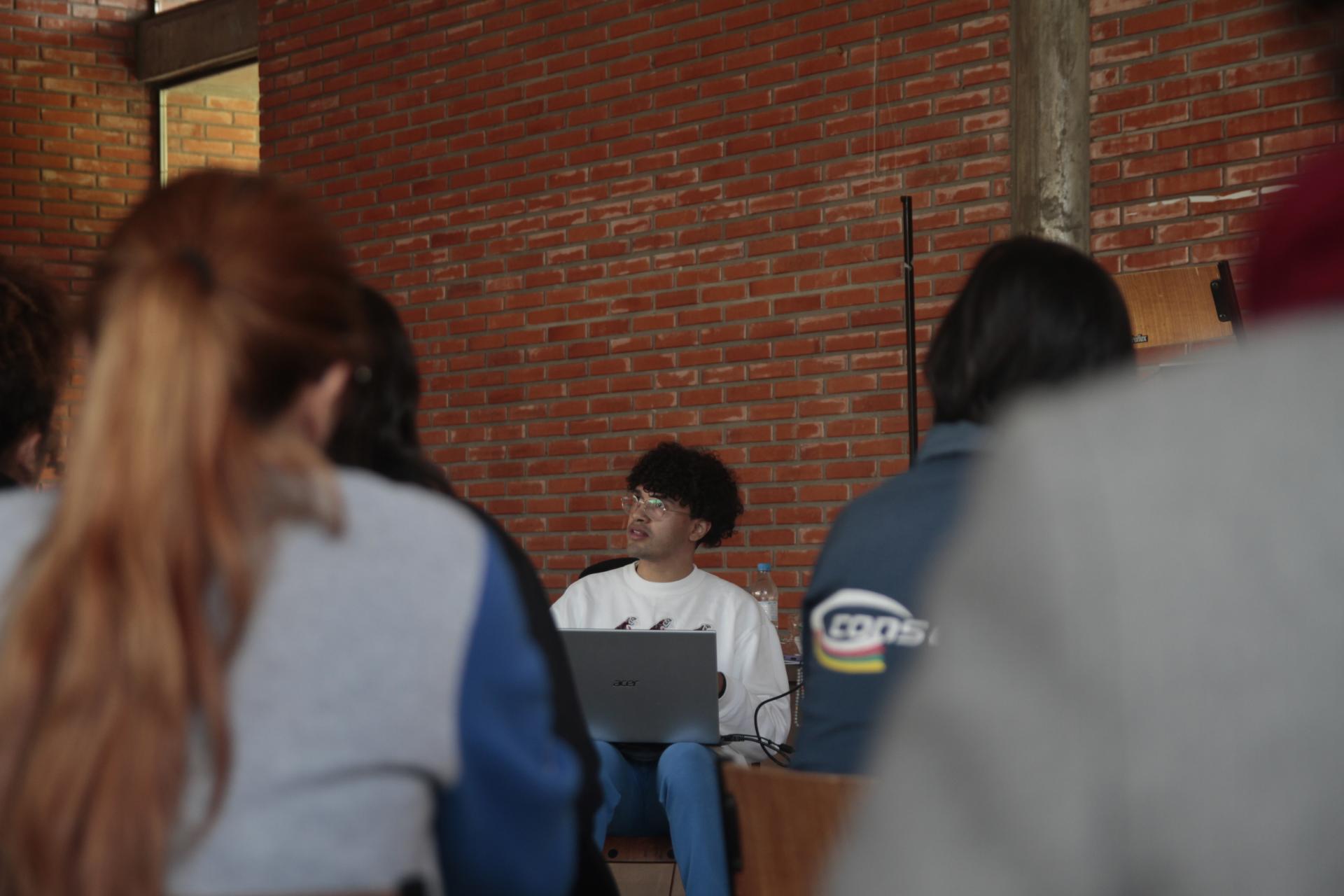 foto colorida horizontal com pessoas sentadas, de costas, voltadas para um rapaz sentado à frente, de moletom branco e com um notebook nas pernas