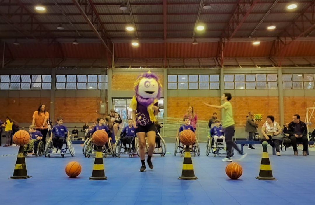 foto colorida horizontal de um ginásio interno com cadeirantes participando de uma atividade com bolas de basquete e cones de trânsito no chão, e ao centro, uma mascote com uma grande máscara na cabeça