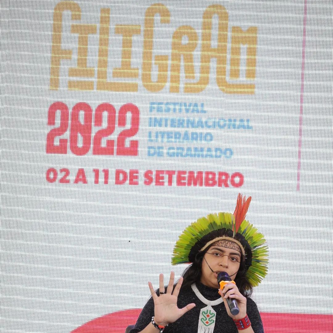 foto colorida quadrada com um grande banner da filigram 2022 ao fundo e abaixo Rayane, de cocar e pintura indígena, falando ao microfone e com a mão direita aberta