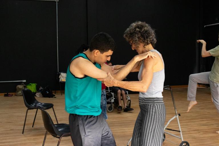 Fotografia horizontal e colorida de Braian Engerrof e sua mãe Maria Iria Engerroff na aula de Dança. Eles estão enquadrados dos joelhos para cima, em pé, de frente um para outro, de lado para nós. Braian é um jovem branco, magro, de estatura média e de cabelos pretos. Ele está vestindo uma regata esverdeada uma bermuda cinza escura. Maria Iria é uma mulher branca, de estatura média, magra, de cabelo curto, cacheado e castanho claro. Ela está vestindo uma regata cinza e uma calça cinza com listras verticais pretas. Braian está com as mãos nos ombros de Maria Iria enquanto ela segura os cotovelos dele. Eles estão em um ambiente interno, com paredes pretas e chão de madeira marrom.