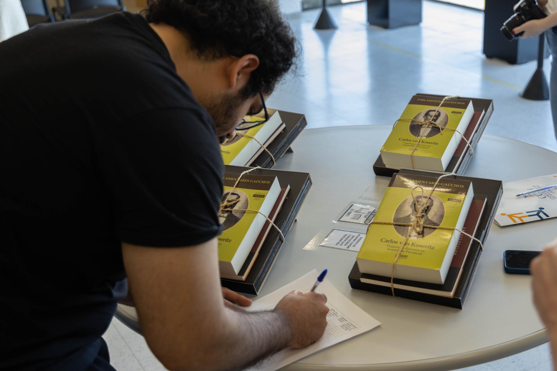 foto colorida retangular de um homem, meio de lado e de costas, camiseta preta, assinando um papel um uma mesa onde estão kits de livros de capa amarela