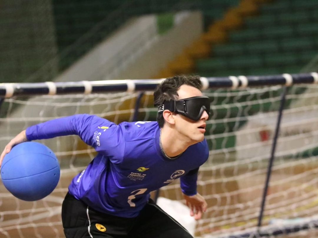 Na fotografia horizontal e colorida, com um chão de madeira e uma goleira ao fundo, o atleta Daniel Gelsdorf está de pé, em posição de ataque, segurando a bola de goalball com a mão direita. Daniel é um homem branco, alto, magro, de cablo curto e castanho escuro, vestindo um óculos preto, o uniforme da equipe da UFSM, que é uma camiseta de manga longa azul escura e uma calça preta.