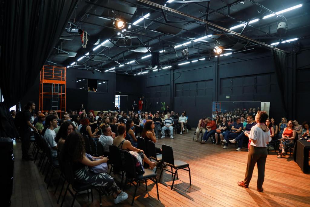 foto colorida horizontal de pessoas sentadas em cadeiras no teatro, de frente e dos lados de uma mulher que está em pé