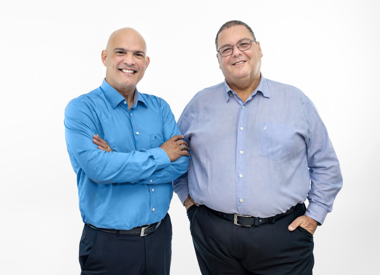 Foto colorida horizontal de dois homens em pé e vestidos com camisa social em tons diferentes de azul e calça social