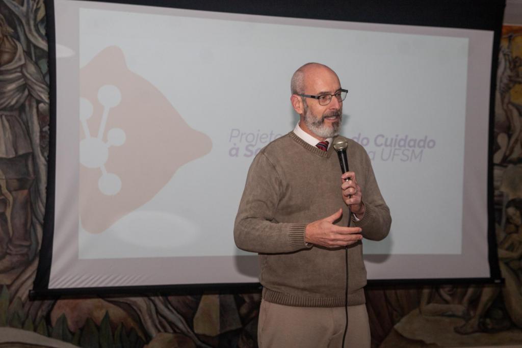 Foto colorida horizontal, ao centro o reitor Luciano Schuch segurando um microfone perto da boca com a mão esquerda. Ao fundo, encoberto, um telão com a logo do projeto.