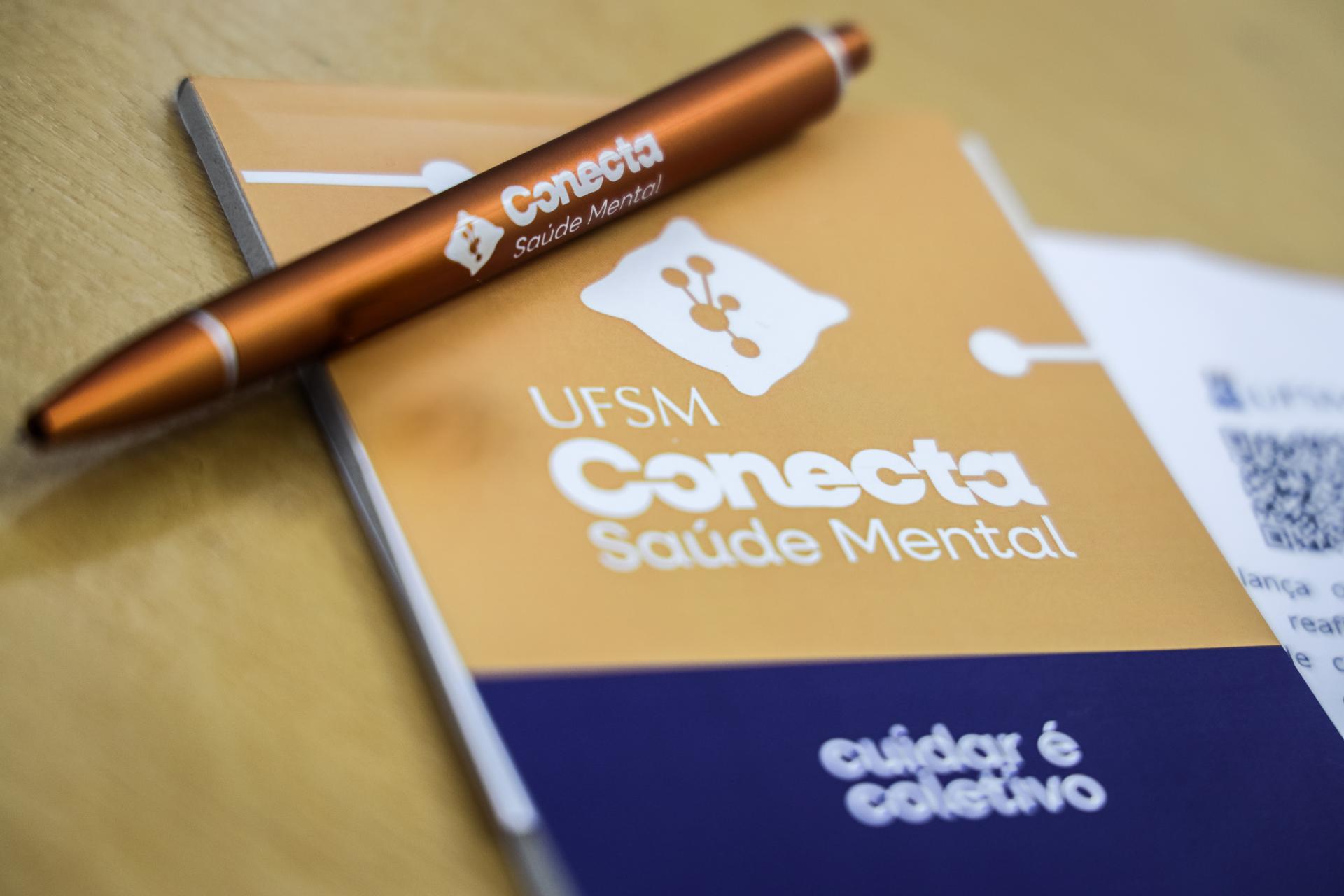 Foto colorida horizontal, da esquerda para o centro uma caneta alaranjada com a logo branca "Conecta Saúde Mental", apoiada sobre um bloco de notas com a logo focada ao centro. Mais abaixo, a frase "Cuidar é coletivo" desfocada.