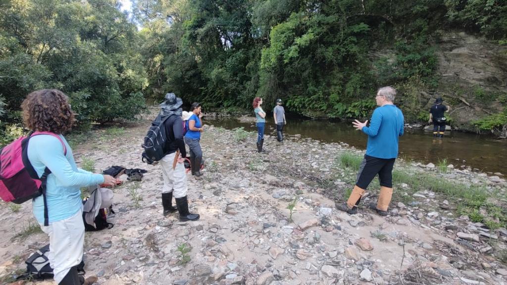 Foto colorida horizontal de grupo de pessoas em um terreno com pedrinhas no chão e vegetação no entorno