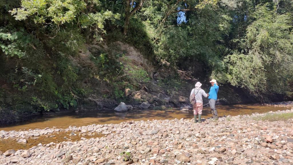 Foto colorida horizontal de uma dois homens ao longe, em frente a um riacho. Ao fundo, um rochedo, que contém os minerais.