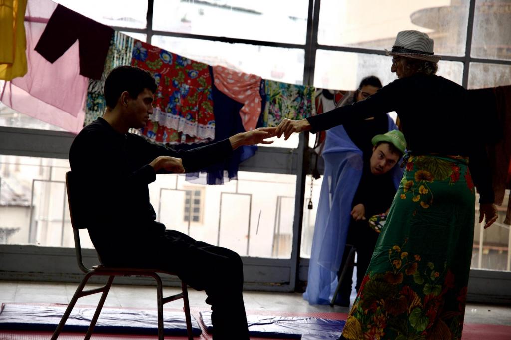 Foto colorida horizontal. Do lado direito, uma mulher idosa segura as mãos de um homem jovem sentado em uma cadeira. Atrás deles, uma varal colorido.