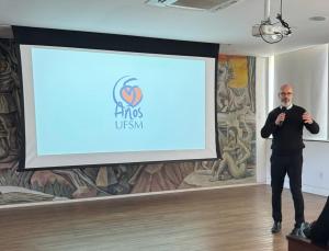 Foto colorida com um telão ao fundo com o selo dos 65 anos e, à frente, o reitor