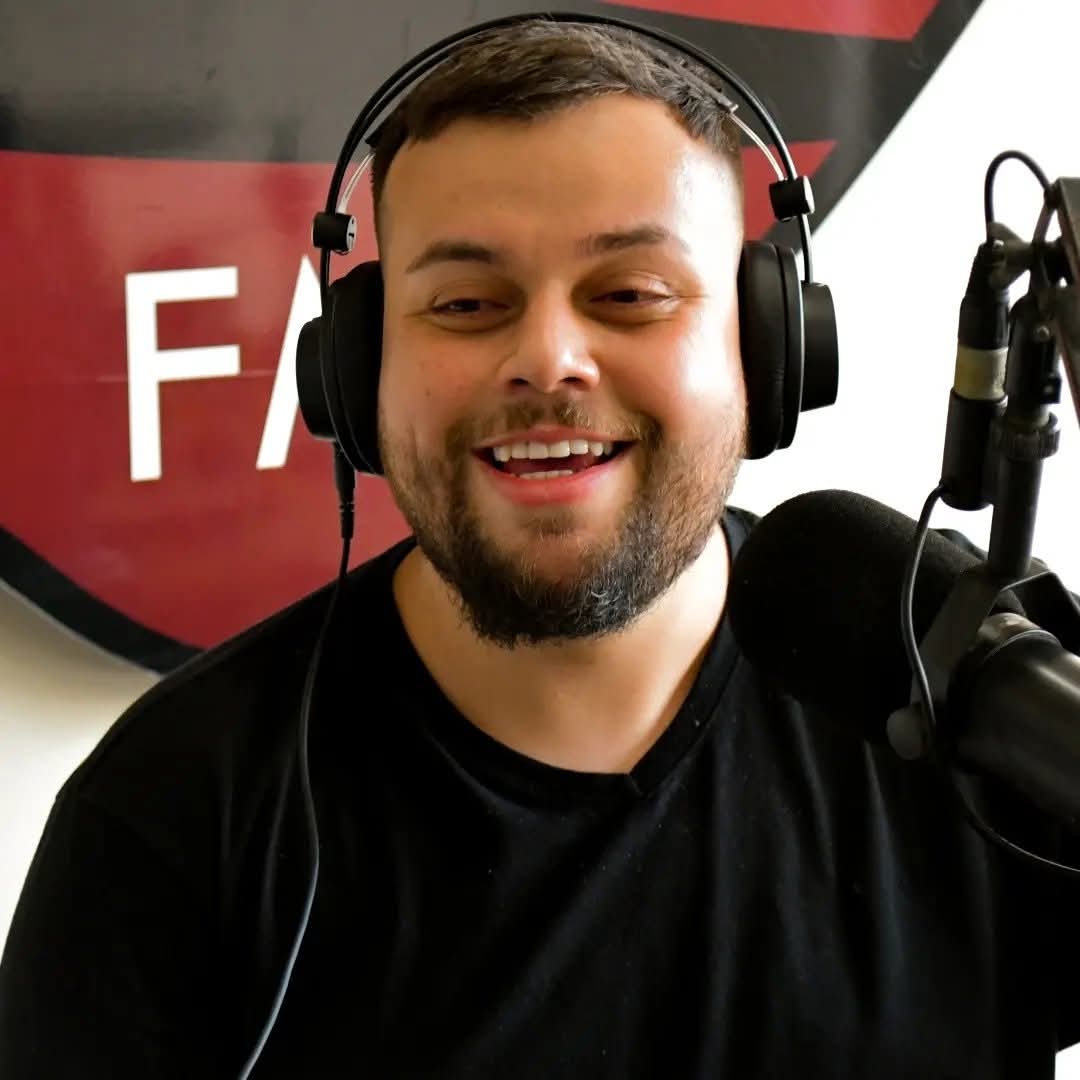 Foto colorida vertical de homem jovem branco com fones de ouvido em frente a um microfone. Ele está sorrindo.