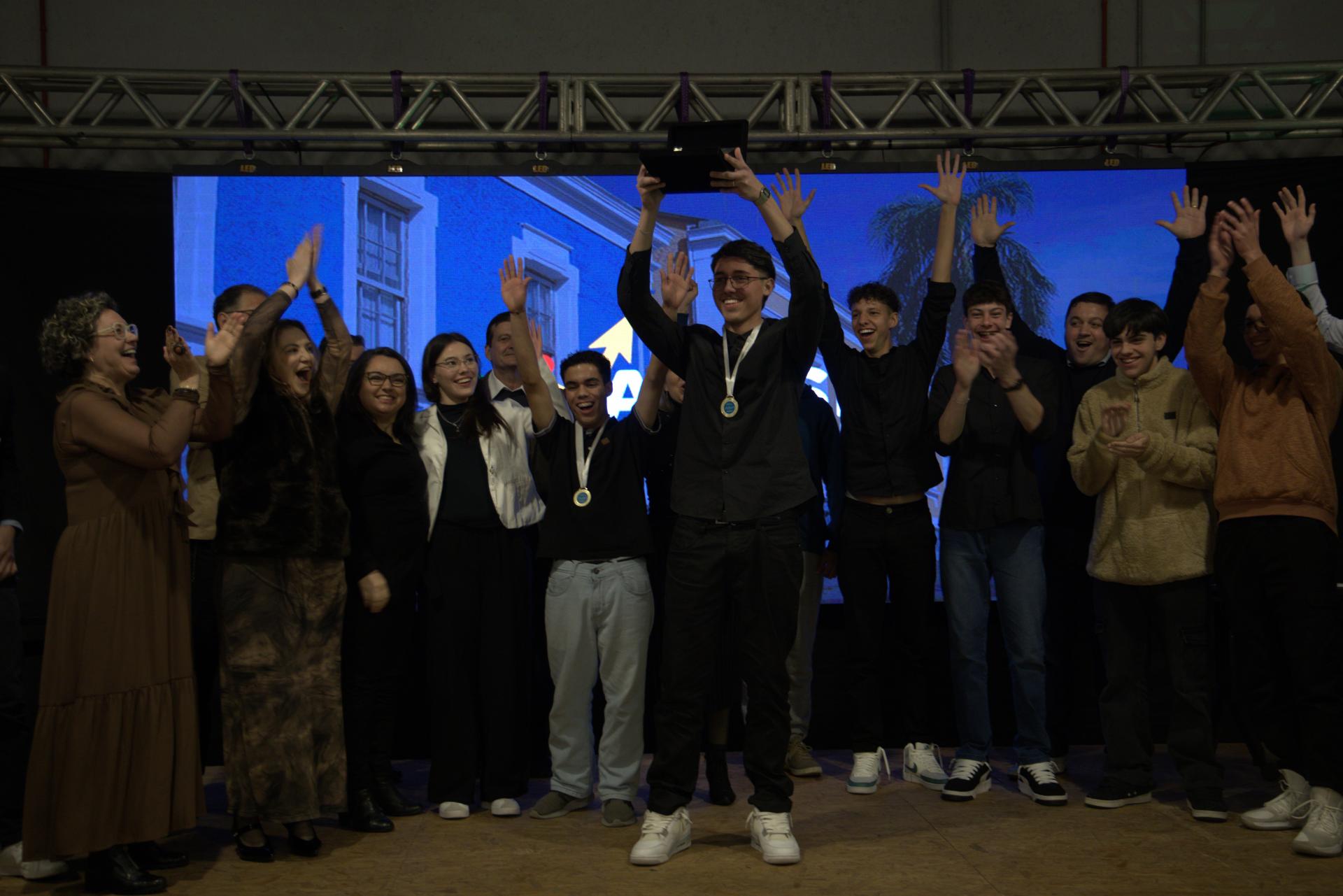 foto colorida horizontal de jovens em pé comemorando em um palco escuro iluminado por um telão atrás. Um deles segura um troféu com as duas mãos acima da cabeça