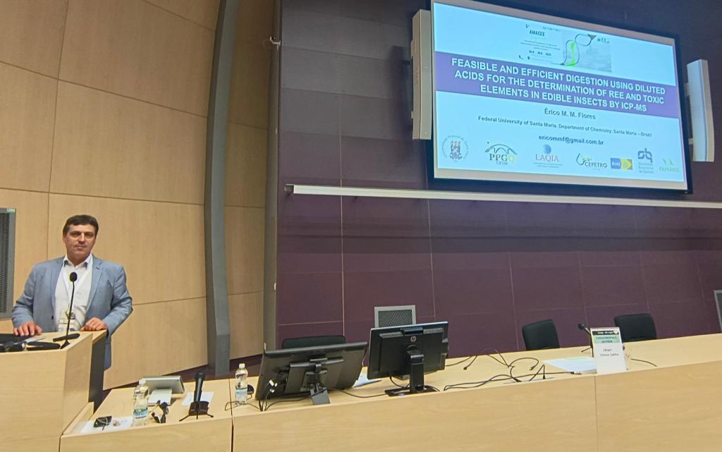 Foto horizontal colorida. A esquerda professor Érico fala em um púlpito. Acima e a direita um telão com imagens relacionadas à pesquisa apresentada