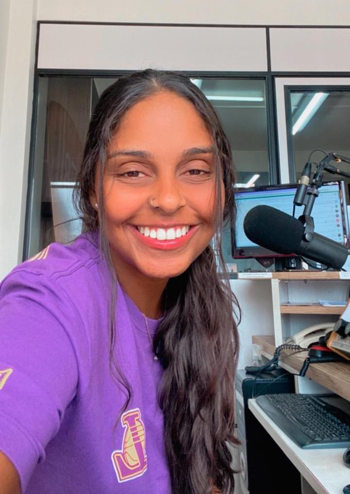 Foto colorida vertical de mulher jovem negra com cabelo comprido. Ela sorri e está em frente ao microfone em um estúdio de rádio