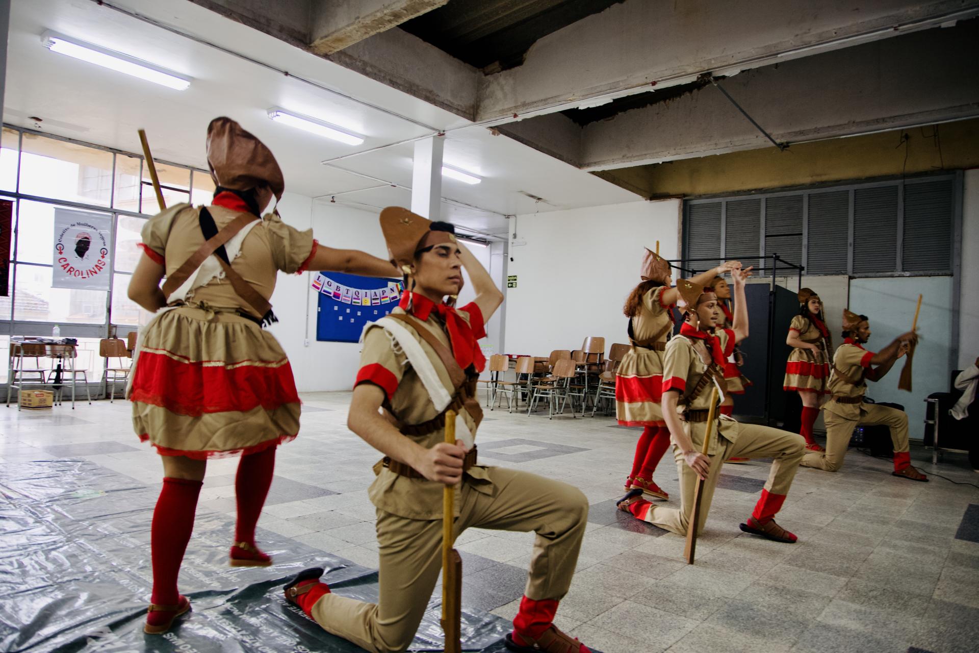 Foto colorida horizontal de seis pessoas vestidas com roupas de cangaceiros nas cores bege, vermelho e branco. Os grupo, em coreografia, conta com três mulheres e três homens. Os homens então com a perna esquerda ajoelhada e as mulheres movimentam-se pelas costas.