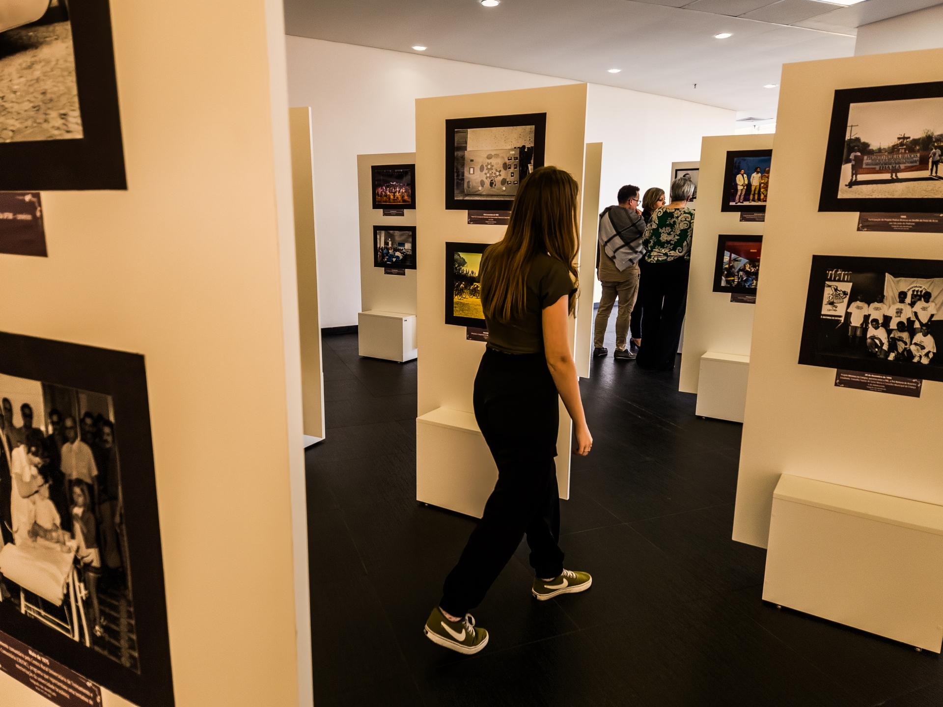 foto colorida horizontal de uma moça de roupa preta caminhando entre armações brancas nas quais estão pendurados quadros em um ambiente interno. Ao fundo há mais pessoas