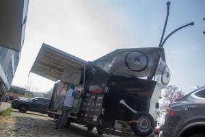 Foto horizontal que mostra um trailer rebocado por um veículo, ambos de cor predominantemente preta. Um dos integrantes do projeto (vestindo boné verde, calças cinza e um colete branco sobre uma camiseta azul) encontra-se de costas sob um dos painéis laterais do trailer.